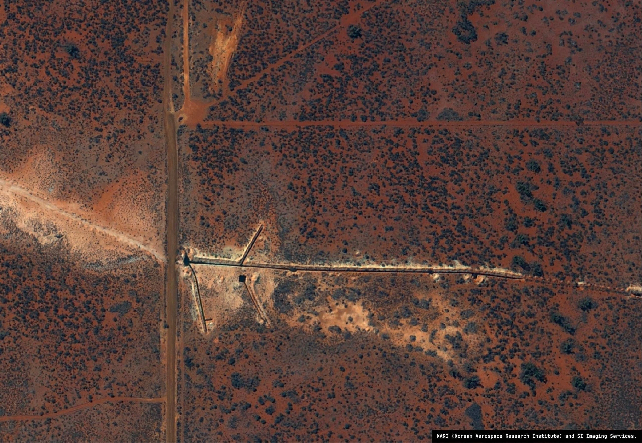 High-resolution satellite imagery of a straight unsealed road through red-soil scrub, with cleared lines and linear earthworks running off it